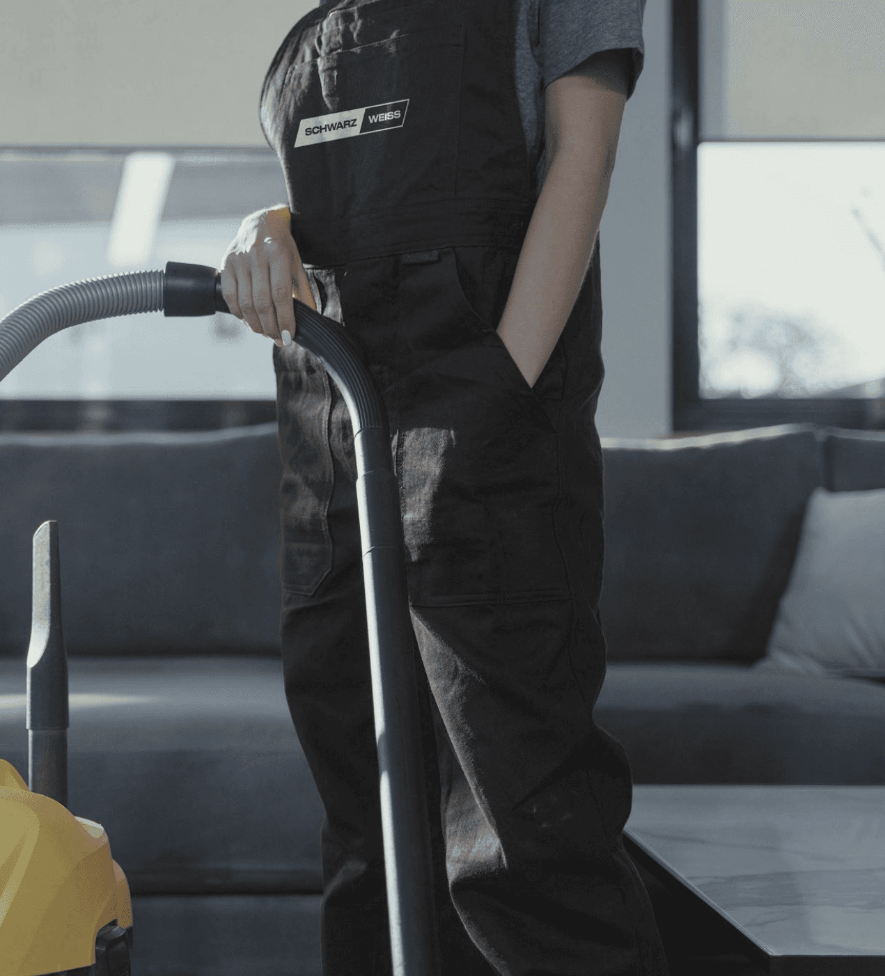 Person in schwarzer Latzhose hält einen Staubsaugerschlauch in einem Wohnzimmer vor einem grauen Sofa.